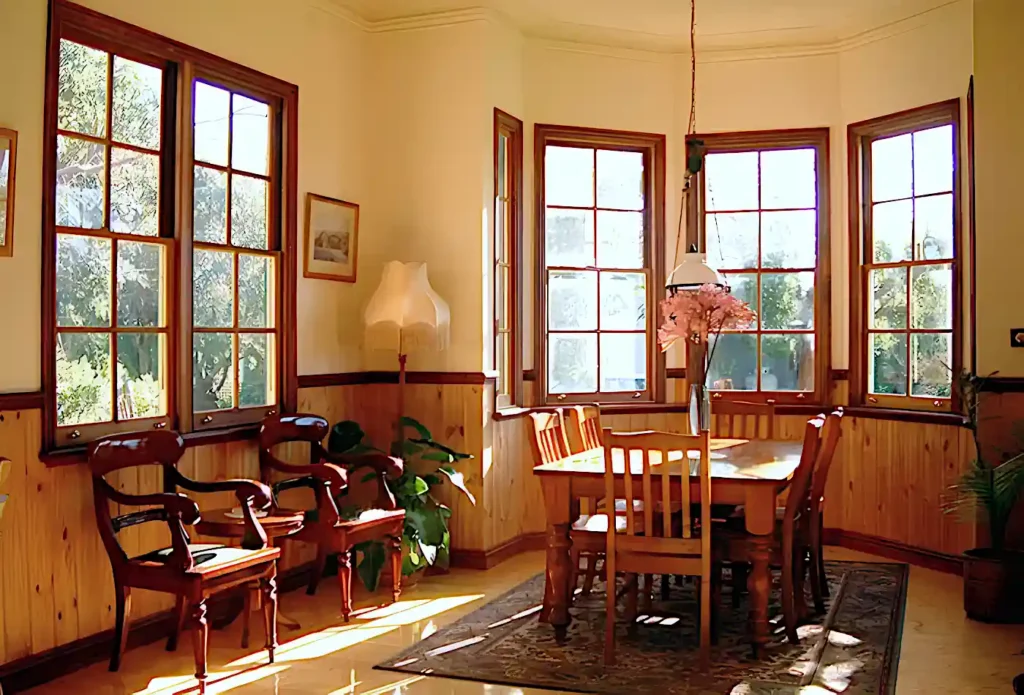 The breakfast area of Butterfly Farm bed and breakfast, with large east and south-facing windows filling the room with warm light. A vase of flowers sits on a large wooden table surrounded by wooden chairs, while two ornate chairs are arranged under a side window. A standard lamp with tasselled shade occupies one corner and sunlight streams through athgelarge bay window behind the table.