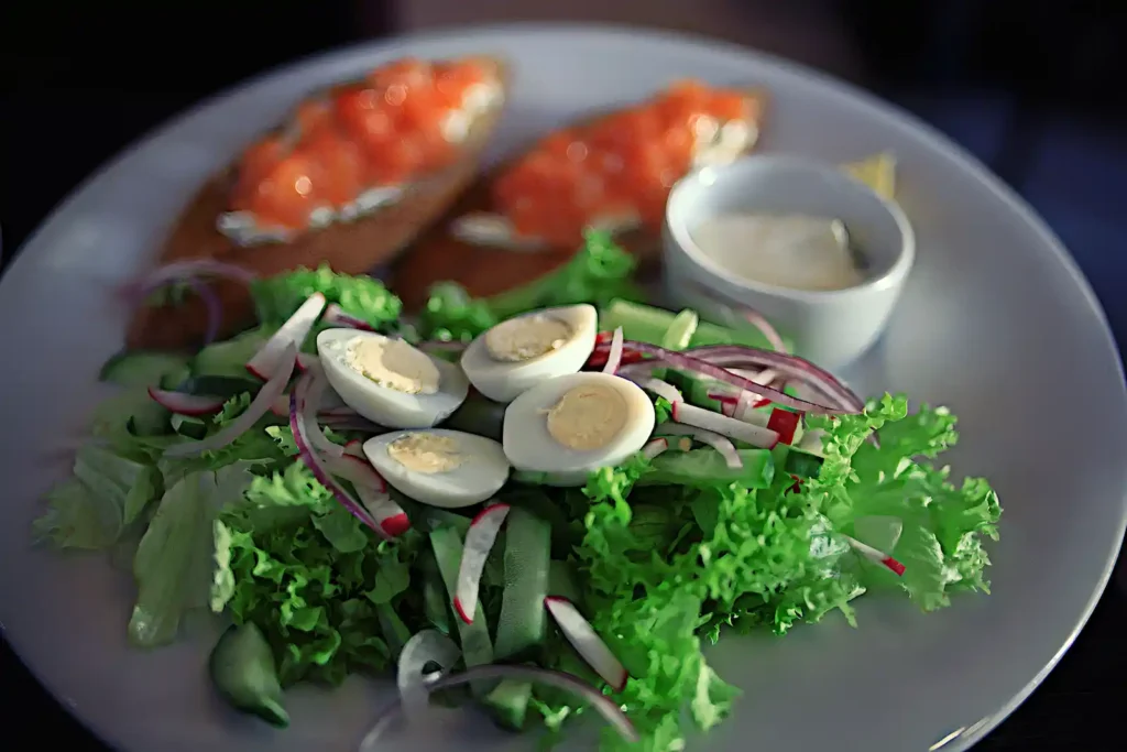 A salad of lettuce cucumber and spanish onions topped with halved boiled eggs sits on a plate with two slices of bruschetta and a small bowl of mayonnaise.   